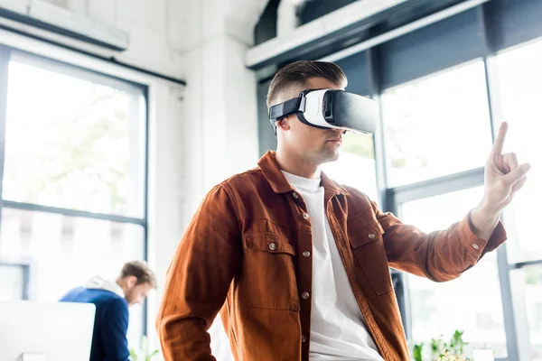 Joven hombre de negocios tocando algo con el dedo mientras usa auriculares de realidad virtual en la oficina - foto de stock