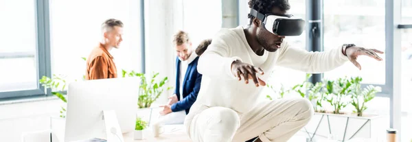 Plano panorámico del hombre de negocios afroamericano usando auriculares vr y sentadilla en el escritorio mientras sus colegas trabajan en la oficina - foto de stock