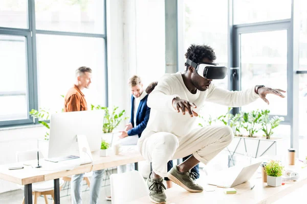 African american businessman squatting on table while using vr headset, and colleagues working in office — Stock Photo