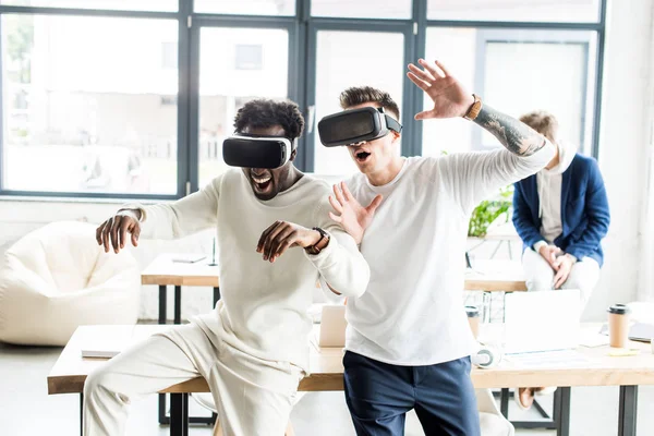 Dos alegres colegas multiculturales haciendo gestos mientras usan auriculares vr en la oficina - foto de stock