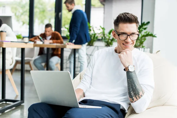 Jovem empresário em óculos usando laptop e sorrindo para a câmera perto de colegas que trabalham no escritório — Fotografia de Stock