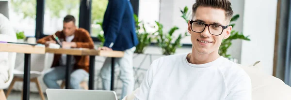 Panoramaaufnahme eines jungen Geschäftsmannes in Brille mit Laptop und lächelnd in die Kamera neben Kollegen, die im Büro arbeiten — Stockfoto