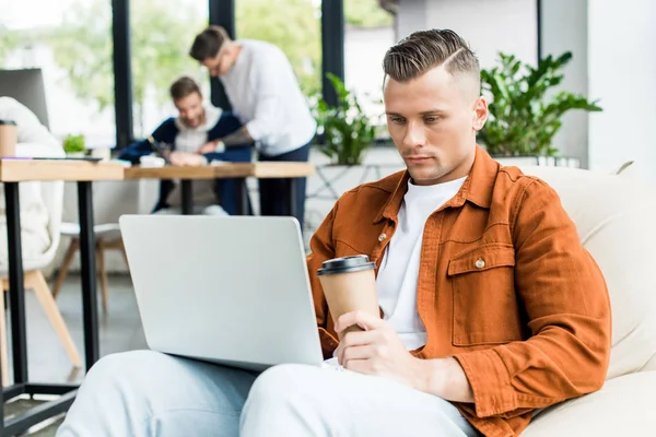 Young businessman holding paper cup and using laptop while working in office near multicultural colleagues — Stock Photo