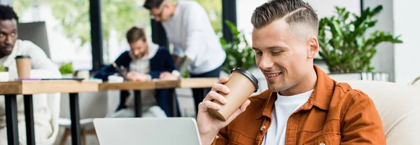 Tiro panorâmico de jovem empresário beber café para ir e usar laptop enquanto trabalhava no escritório perto de colegas multiculturais — Fotografia de Stock