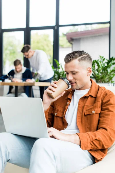 Joven hombre de negocios beber café para ir y el uso de la computadora portátil mientras trabaja en la oficina cerca de colegas - foto de stock