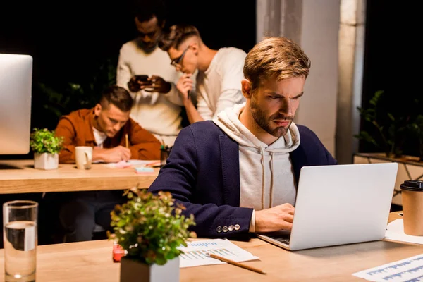 Молодой, серьезный бизнесмен, работающий ночью на ноутбуке рядом с коллегами по мультикультурному бизнесу в офисе — стоковое фото