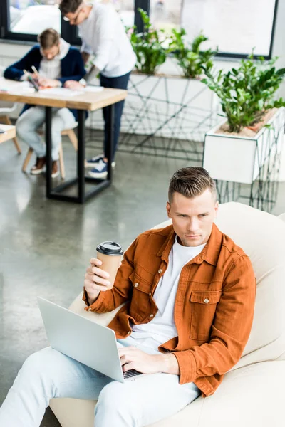 Високий кут зору молодого бізнесмена, який тримає паперовий стаканчик і використовує ноутбук під час роботи в офісі поблизу колег — стокове фото