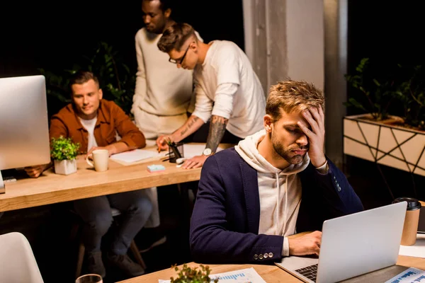 Exhausted businessman using laptop while working in office at night near multicultural colleagues — Stock Photo