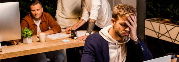 Panoramaaufnahme eines erschöpften Geschäftsmannes mit Laptop während seiner nächtlichen Arbeit im Büro in der Nähe von Kollegen — Stockfoto