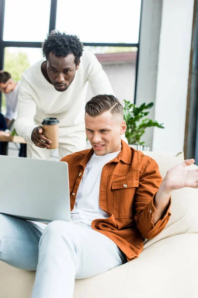 Hombre de negocios afroamericano sosteniendo café para ir y señalando con el dedo a la computadora portátil en manos de colega mostrando gesto encogiéndose de hombros - foto de stock