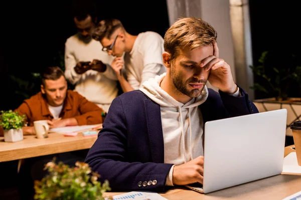 Втомлений бізнесмен використовує ноутбук під час роботи в офісі вночі біля мультикультурних колег — стокове фото