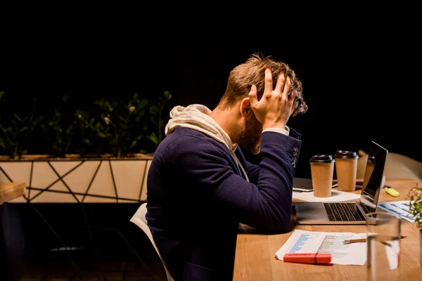Joven hombre de negocios agotado sosteniendo las manos en la cabeza mientras se sienta en la noche en la oficina cerca de la computadora portátil y vasos de papel - foto de stock