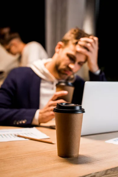 Messa a fuoco selettiva di tazza usa e getta vicino uomo d'affari esausto che lavora di notte in ufficio — Foto stock