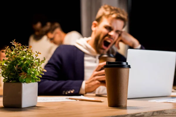 Selective focus of disposable cup near exhausted businessman yawning while working at night in office — Stock Photo