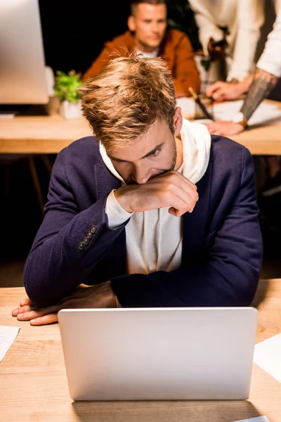 Junger, erschöpfter Geschäftsmann schaut auf Laptop, während er nachts im Büro in der Nähe von Kollegen arbeitet — Stockfoto