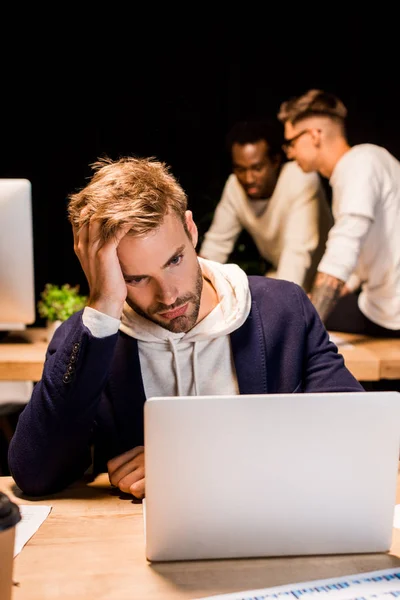 Homem de negócios cansado olhando para laptop enquanto trabalhava à noite no escritório perto de colegas multiculturais — Fotografia de Stock