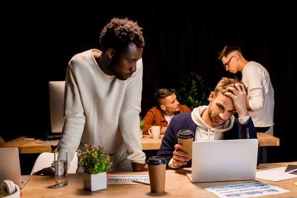 Afrikanisch-amerikanischer Geschäftsmann steht neben erschöpftem Kollegen und hält Kaffee to go, während er nachts im Büro arbeitet — Stockfoto