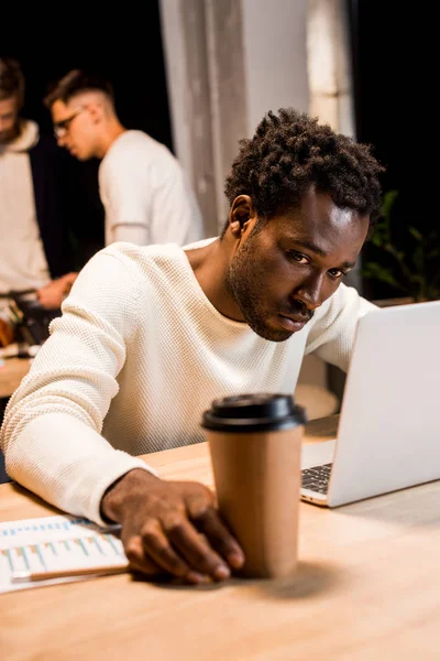 Избирательный фокус уставшего африканского американского бизнесмена, держащего кофе, чтобы пойти во время работы ночью в офисе рядом с коллегами — стоковое фото