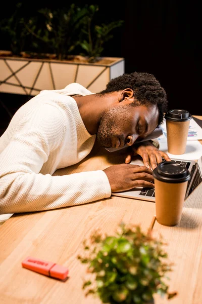 Stanco uomo d'affari africano americano che dorme sul posto di lavoro vicino al computer portatile e tazze usa e getta di notte in ufficio — Foto stock