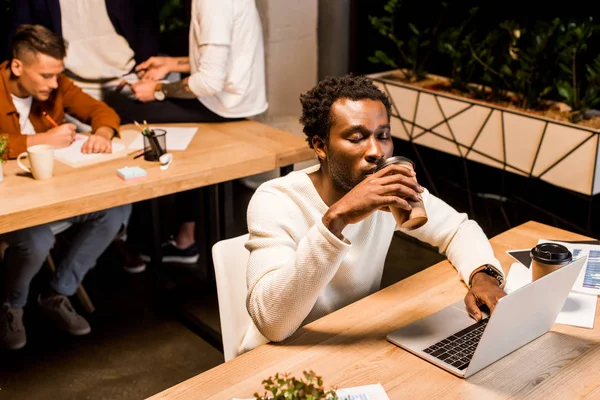 Müder afrikanisch-amerikanischer Geschäftsmann trinkt Kaffee to go, während er nachts am Arbeitsplatz in der Nähe von Kollegen im Büro sitzt — Stockfoto