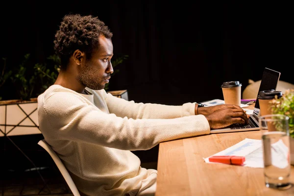 Müder afrikanisch-amerikanischer Geschäftsmann sitzt am Arbeitsplatz neben Laptop und Einwegbechern in der Nacht im Büro — Stockfoto