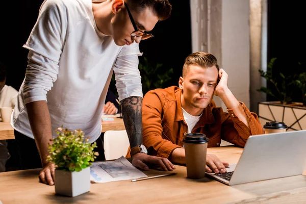 Young businessman standing near exhausted colleague using laptop at night in office — Stock Photo