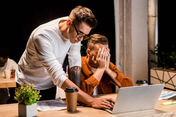 Молодий бізнесмен стоїть біля виснаженого колеги, покриваючи обличчя руками, сидячи біля ноутбука в офісі — стокове фото