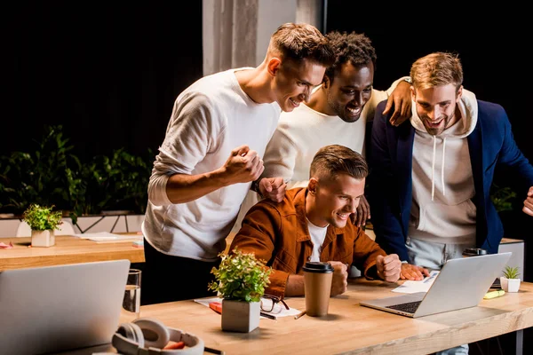 Heureux jeunes hommes d'affaires regardant ordinateur portable et montrant geste gagnant la nuit au bureau — Photo de stock