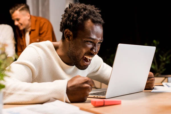 Emocionado hombre de negocios afroamericano mirando a la computadora portátil mientras trabaja en la noche en la oficina - foto de stock