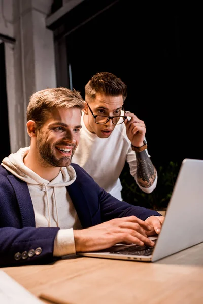 Jovem homem de negócios tocando óculos enquanto olha para laptop perto de colega feliz — Fotografia de Stock