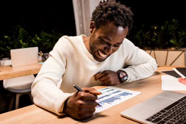 Happy african american businessman looking at graphs and charts while working at night in office — Stock Photo