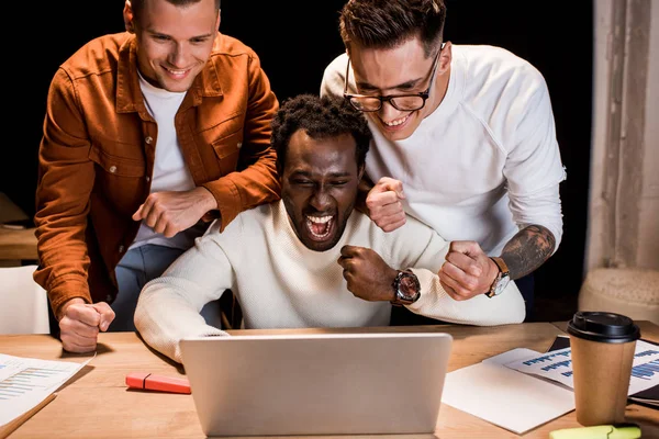 Empresários multiculturais felizes mostrando gesto vencedor enquanto olha para laptop à noite no escritório — Fotografia de Stock