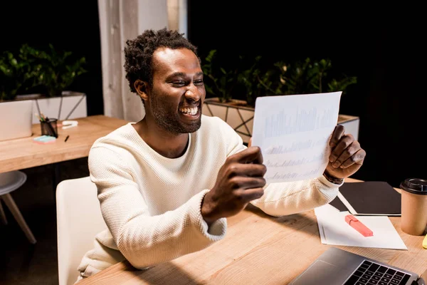 Happy african american businessman looking at paper while working at night in office — Stock Photo