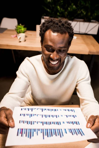 Щасливий афроамериканський бізнесмен тримає папір з графіками і графіками під час роботи вночі в офісі — стокове фото