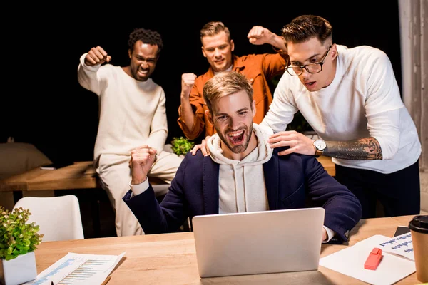 Felices empresarios multiculturales mostrando un gesto ganador mientras trabajan de noche en la oficina - foto de stock