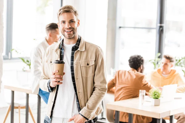 Joven, hombre de negocios guapo sosteniendo café para ir y sonriendo a la cámara - foto de stock