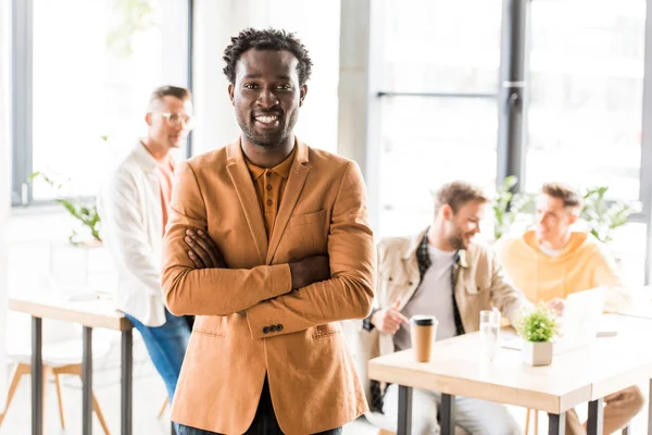 Empresário americano africano alegre de pé com braços cruzados perto de colegas no escritório e sorrindo para a câmera — Fotografia de Stock