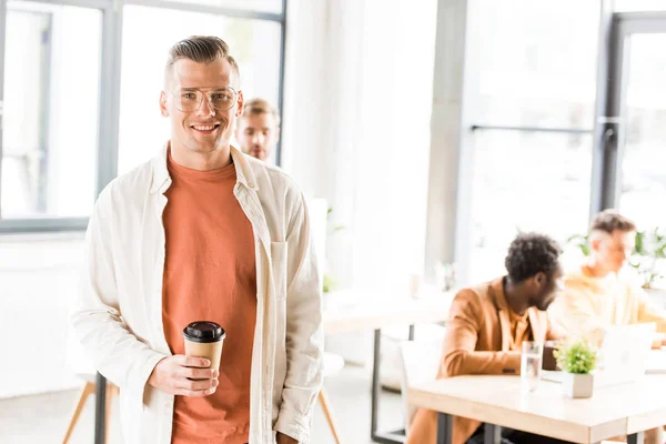 Joven, hombre de negocios guapo sosteniendo café para ir y sonriendo a la cámara - foto de stock