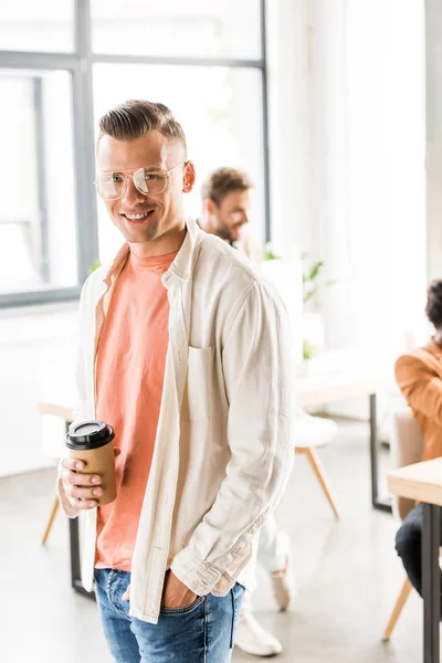 Young businessman standing with hand and pocket, holding coffee to go and smiling at camera — Stock Photo