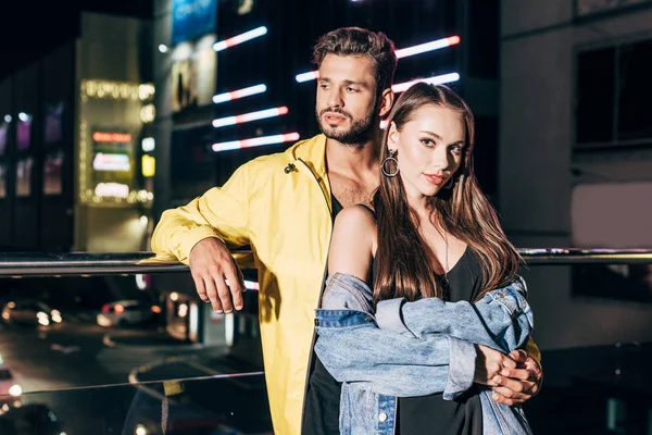 Handsome boyfriend and attractive girlfriend in jacket hugging in night city — Stock Photo