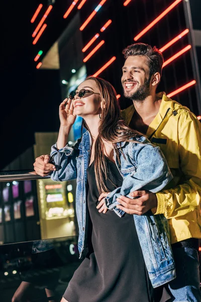 Atractiva novia y novio guapo abrazando y mirando hacia otro lado en la ciudad de la noche - foto de stock