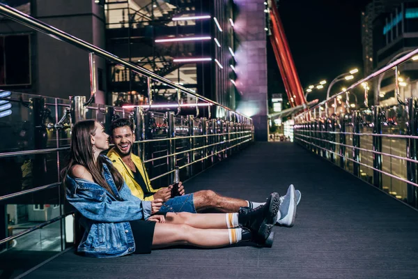 Vista laterale del fidanzato con bottiglia e fidanzata sorridente seduta nella città notturna — Foto stock