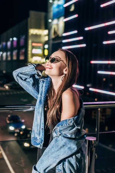 Mujer atractiva en chaqueta de mezclilla y gafas de sol sonriendo y mirando hacia otro lado en la ciudad de la noche - foto de stock