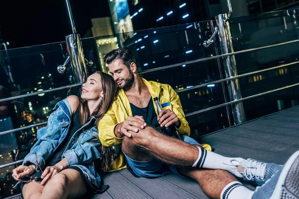 Attractive girlfriend lying on shoulder of her boyfriend with bottle in night city — Stock Photo