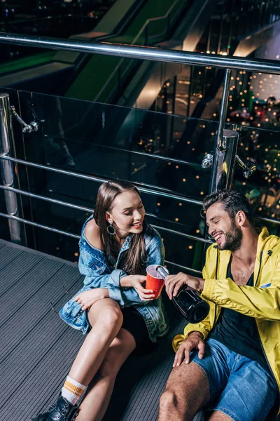 Vue grand angle de beau petit ami souriant versant du champagne à la tasse en plastique dans la ville de nuit — Photo de stock