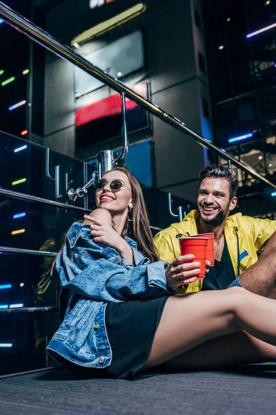 Guapo novio con taza de plástico y atractiva novia señalando con el dedo en la ciudad de la noche - foto de stock