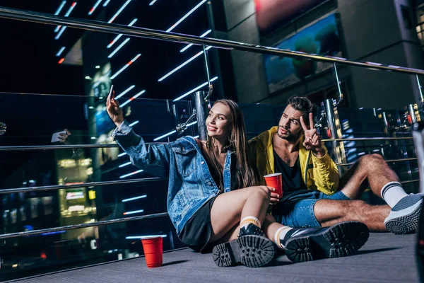 Handsome boyfriend showing peace sign and attractive girlfriend taking selfie in night city — Stock Photo