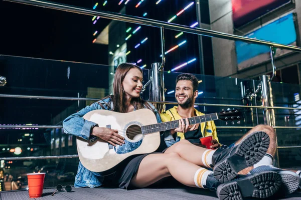 Handsome boyfriend with plastic cup and attractive girlfriend playing acoustic guitar in night city — Stock Photo