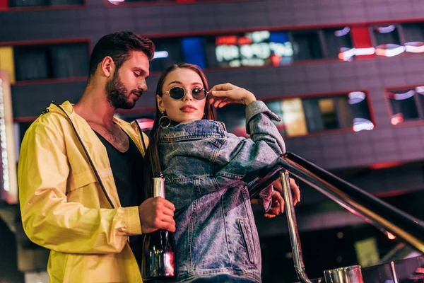 Guapo novio con botella abrazando atractiva novia en chaqueta de mezclilla en la ciudad de noche - foto de stock