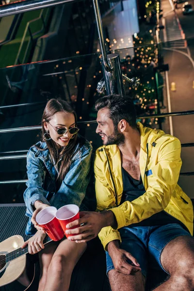 Vue grand angle de beau petit ami et jolie petite amie cliquetis avec des tasses en plastique dans la ville de nuit — Photo de stock